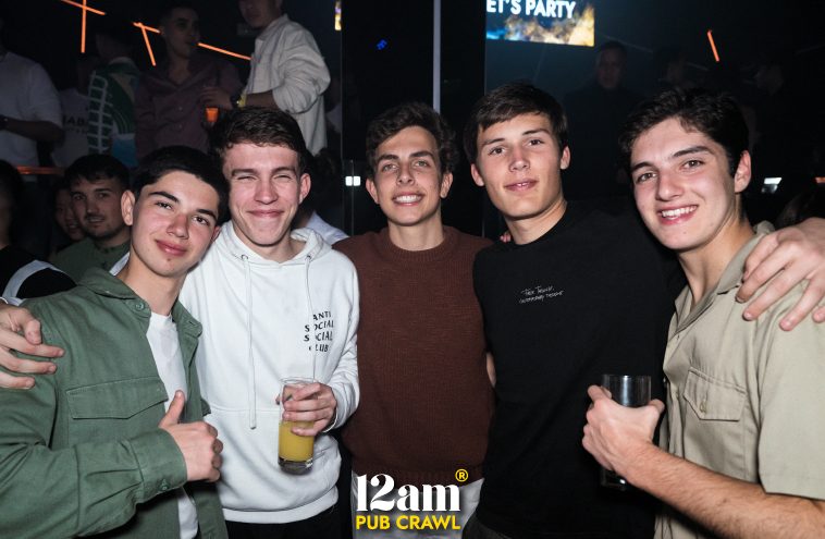 Group of smiling young men, including Erasmus students and solo travelers, enjoying drinks and the lively atmosphere at a Madrid club during a 12am Pub Crawl. Highlighting new friendships and fun on a Madrid night out.