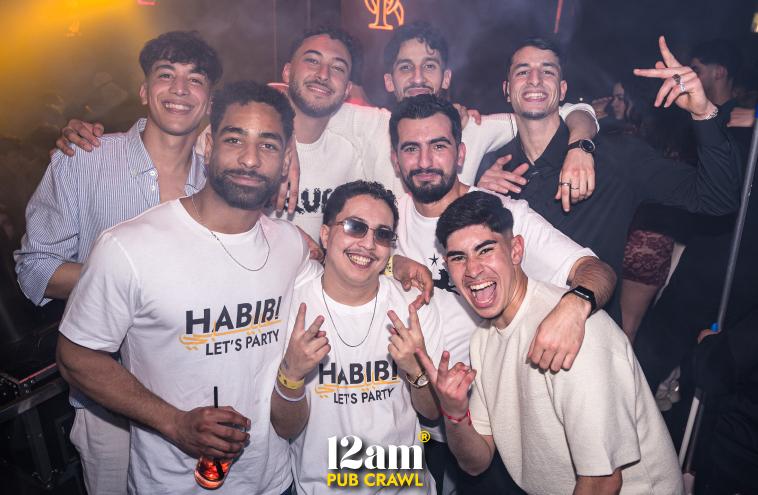 Group of excited young adults, including Erasmus students and solo travelers, posing with 12am Pub Crawl staff wearing "Habibi Let's Party" t-shirts at an Arabian night event in ICON Madrid Club. Showcasing the unique themed nightlife of a Madrid pub crawl.