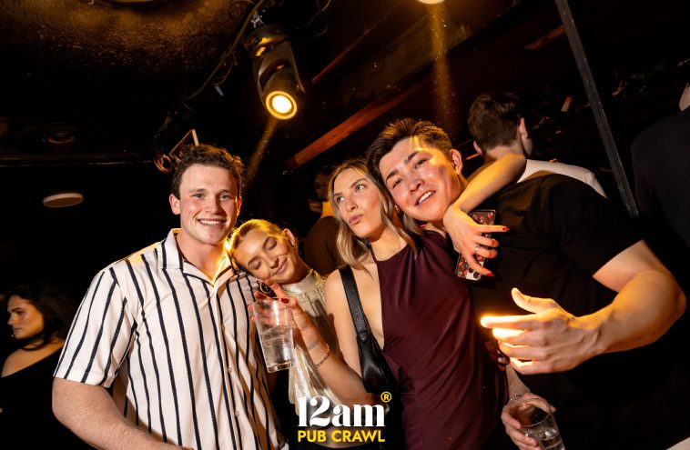Four happy young adults, likely Erasmus students or solo travelers, smiling and enjoying drinks in a vibrant Madrid club during a 12am Pub Crawl. Capturing the fun social atmosphere of a Madrid pub crawl.